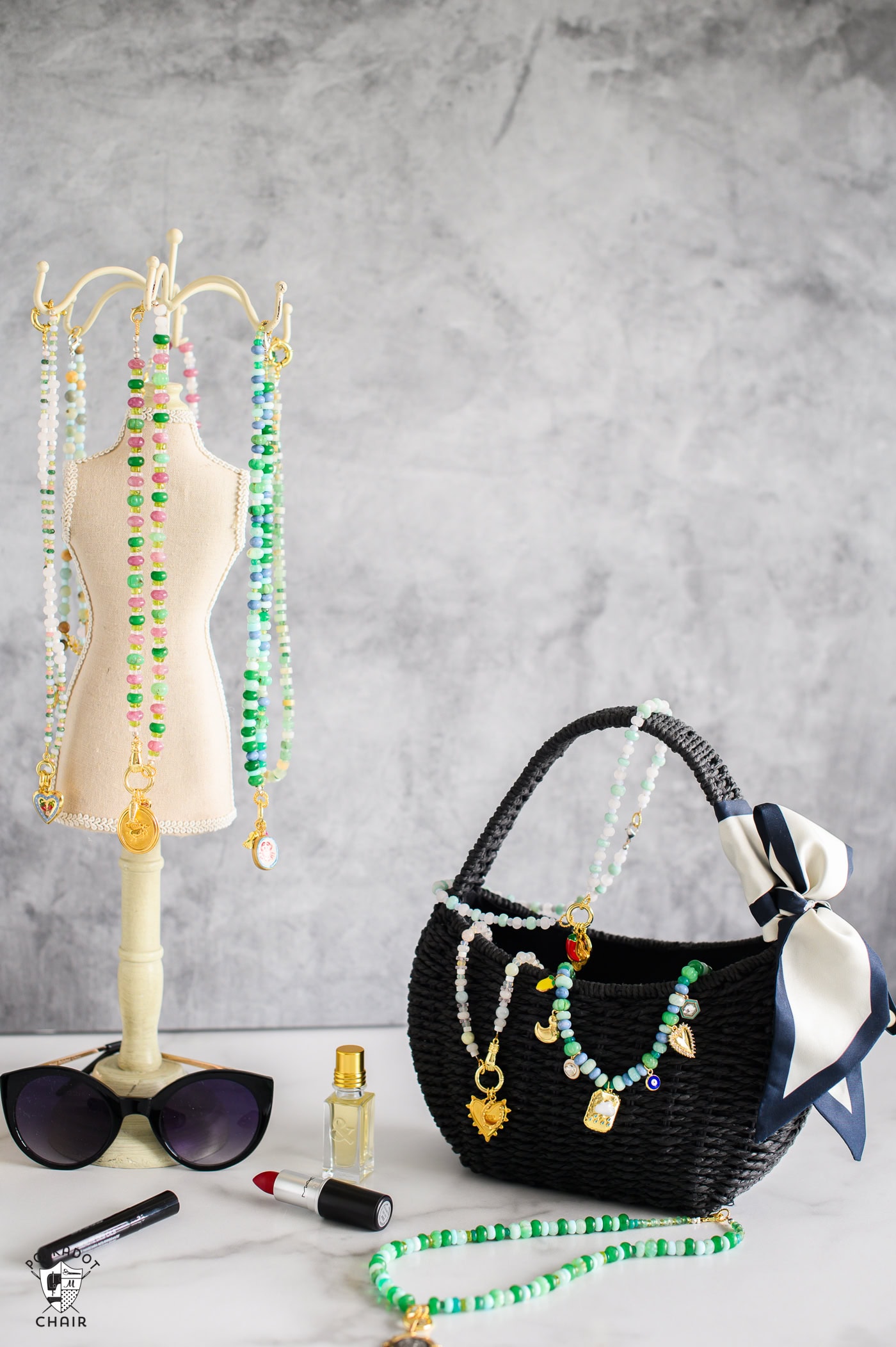 mini mannequin with green and pink charm necklaces hanging from the top with gold charms and a black purse with blue and green necklaces draped across it. sunglasses and lipstick and perfume in front of the table.
