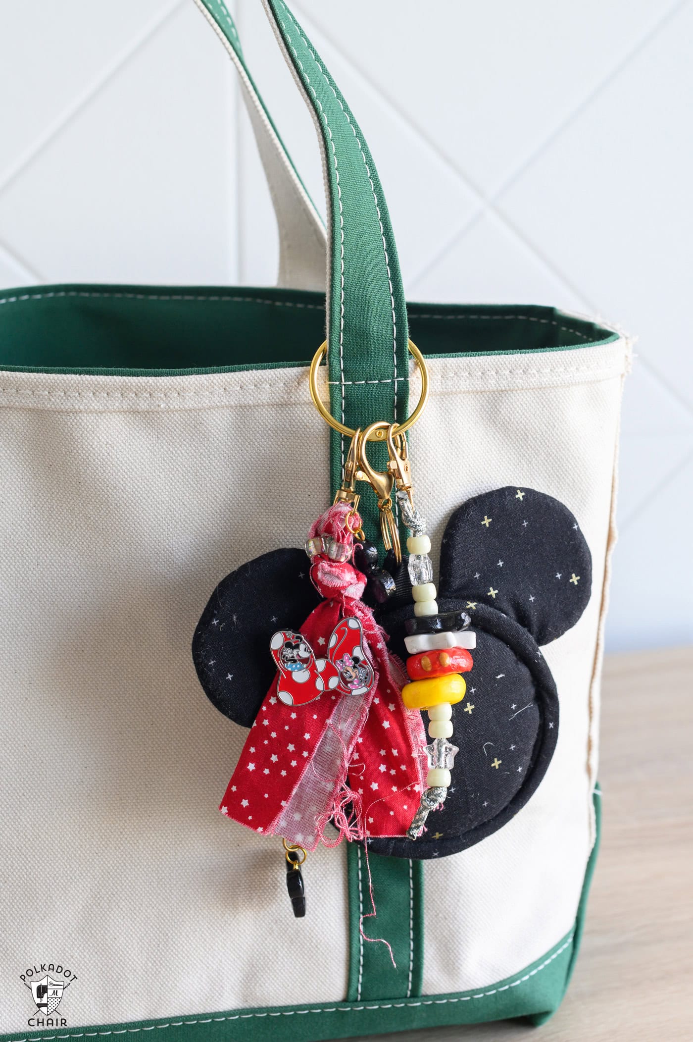 white canvas tote with green straps, with a black mickey head fabric charm. with a red fabric tied to a key chain with a red bow pin and a key chain with white beads and black white, red, yellow clay beads in a stack.