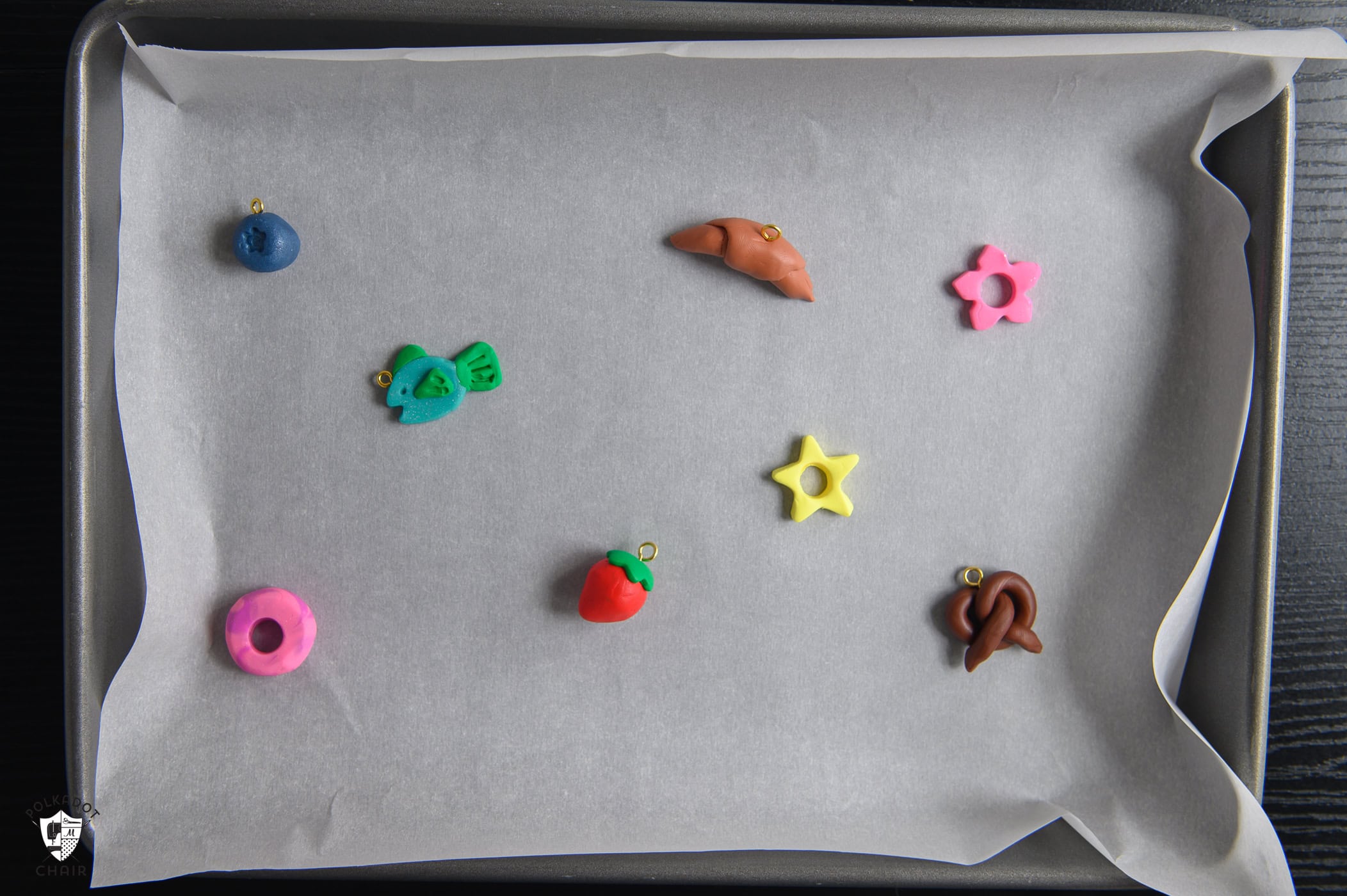 cookie sheet with clay beads and charms spread out in shape of blueberry, croissant, flower, fish, star, strawberry, pretzel, pink swirl
