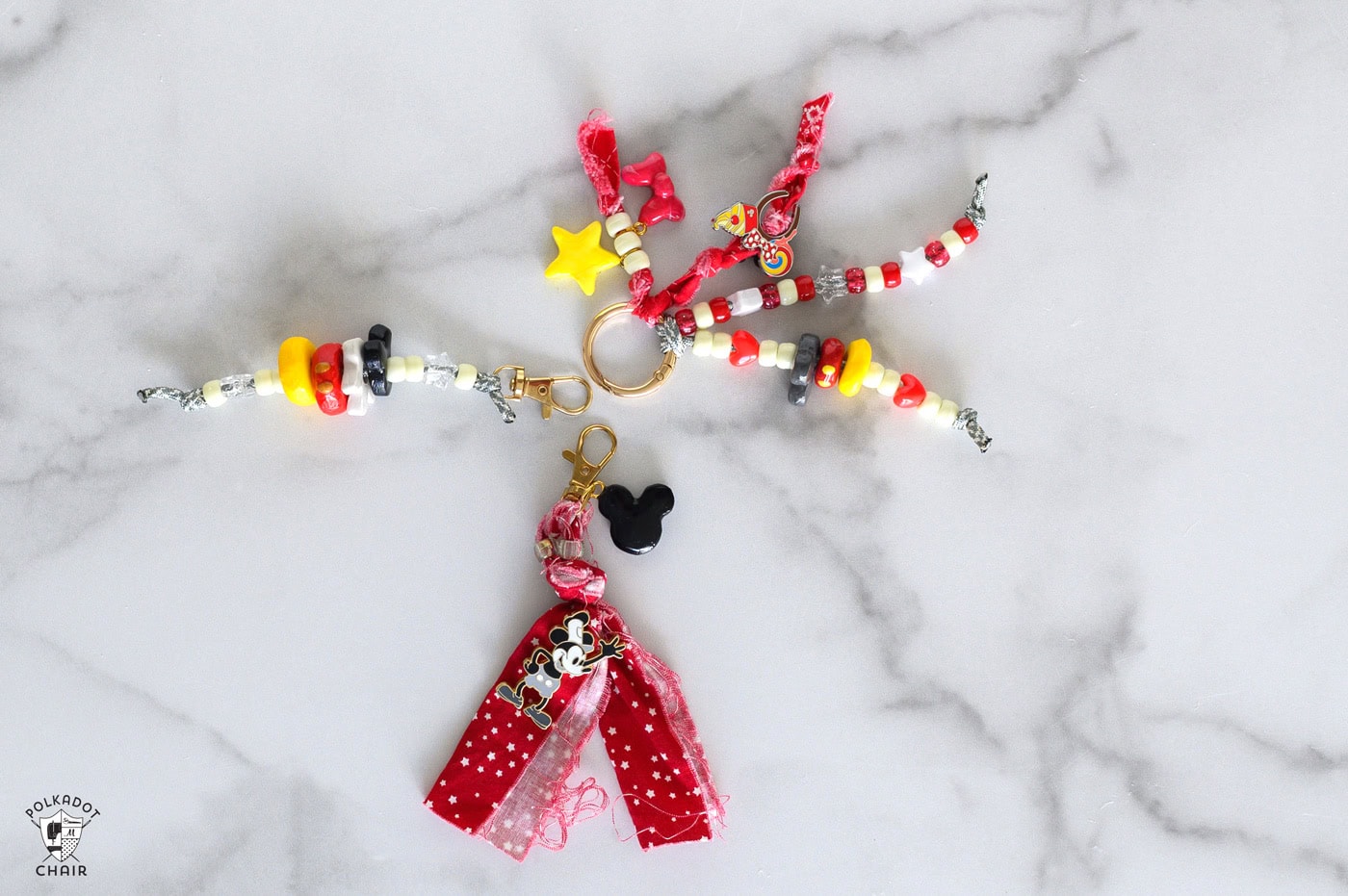 white background with three key chains with white, red, and yellow beads. one with red fabric and a micky mouse pin and charm, one with a black, white, red and yellow clay bead stack, and one with several strands of beads and a red fabric braid.
