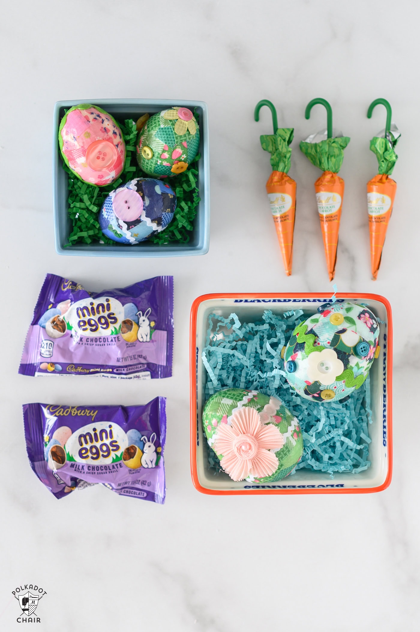 2 berry baskets with blue, green, and pink fabric covered eggs decorated with ricrac and buttons. three chocolate carrots and two mini bags of cadbury mini eggs.