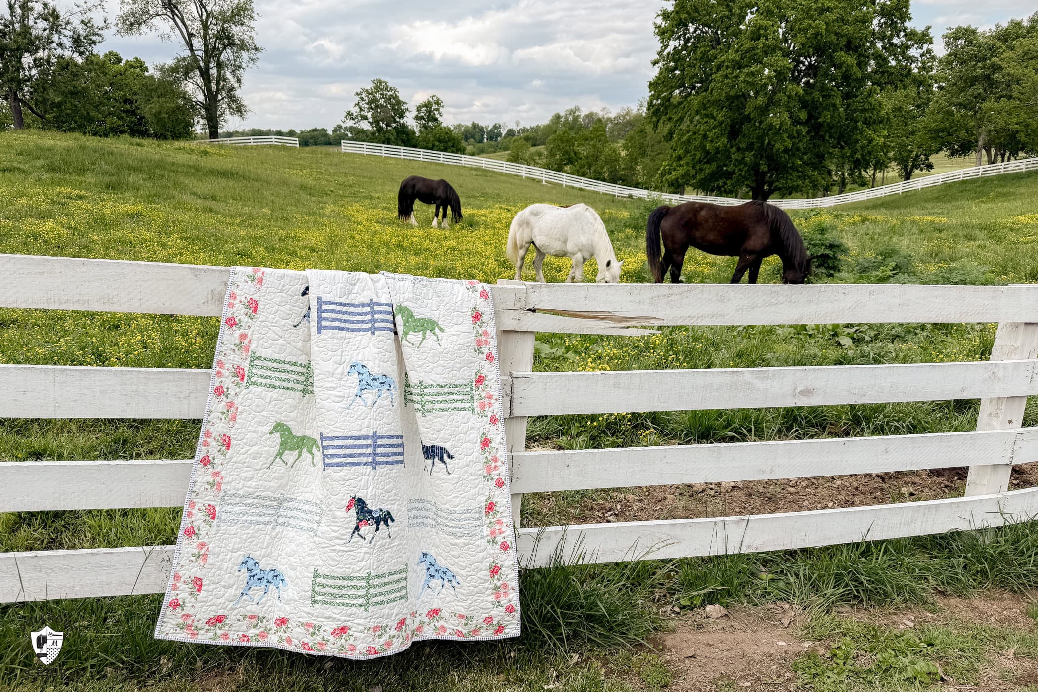 horse quilt hanging on white horse fence in front of field with horses and yellow flowers