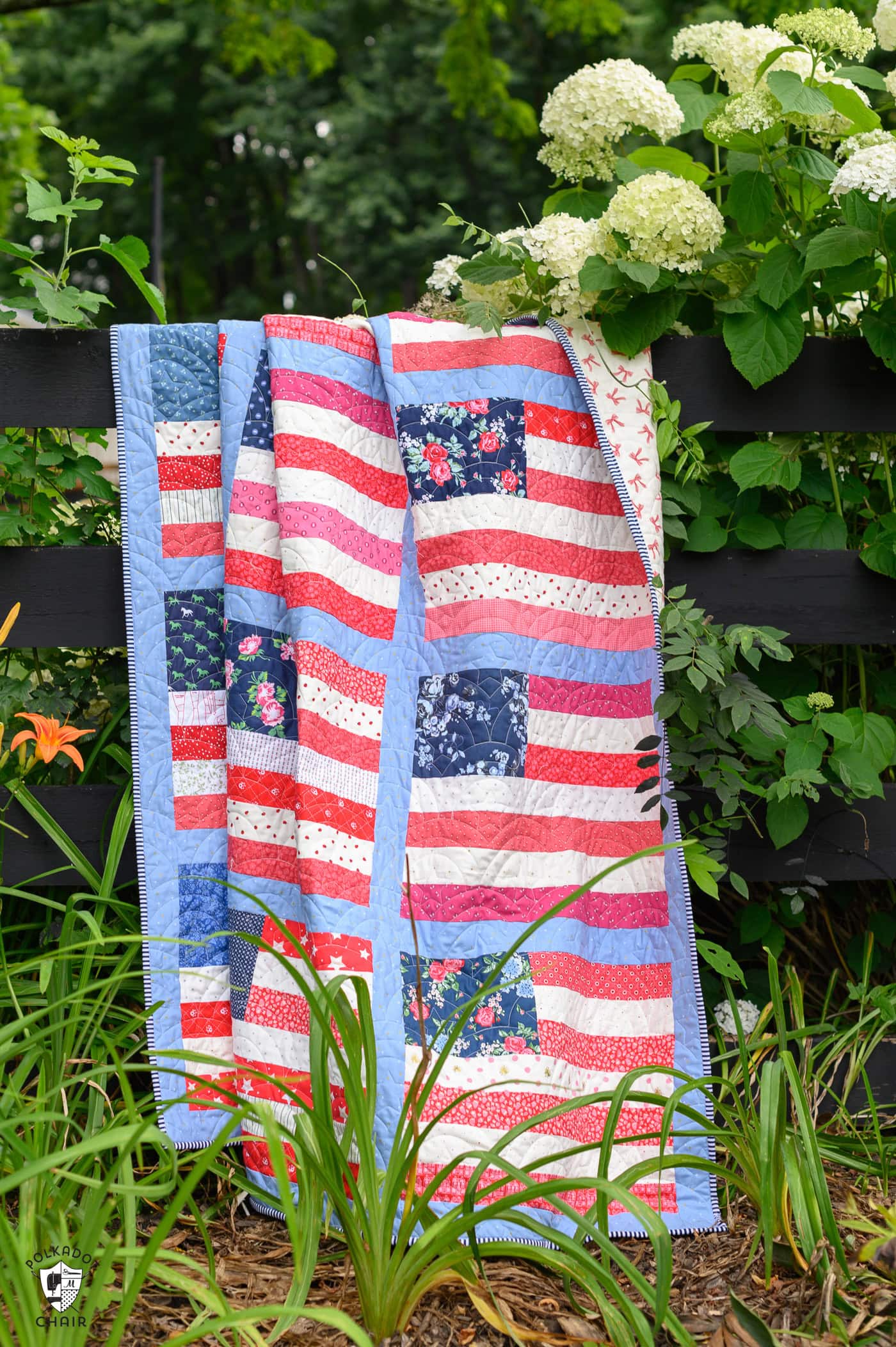 Red, white and blue American flag quilt on fence outdoors with flowers