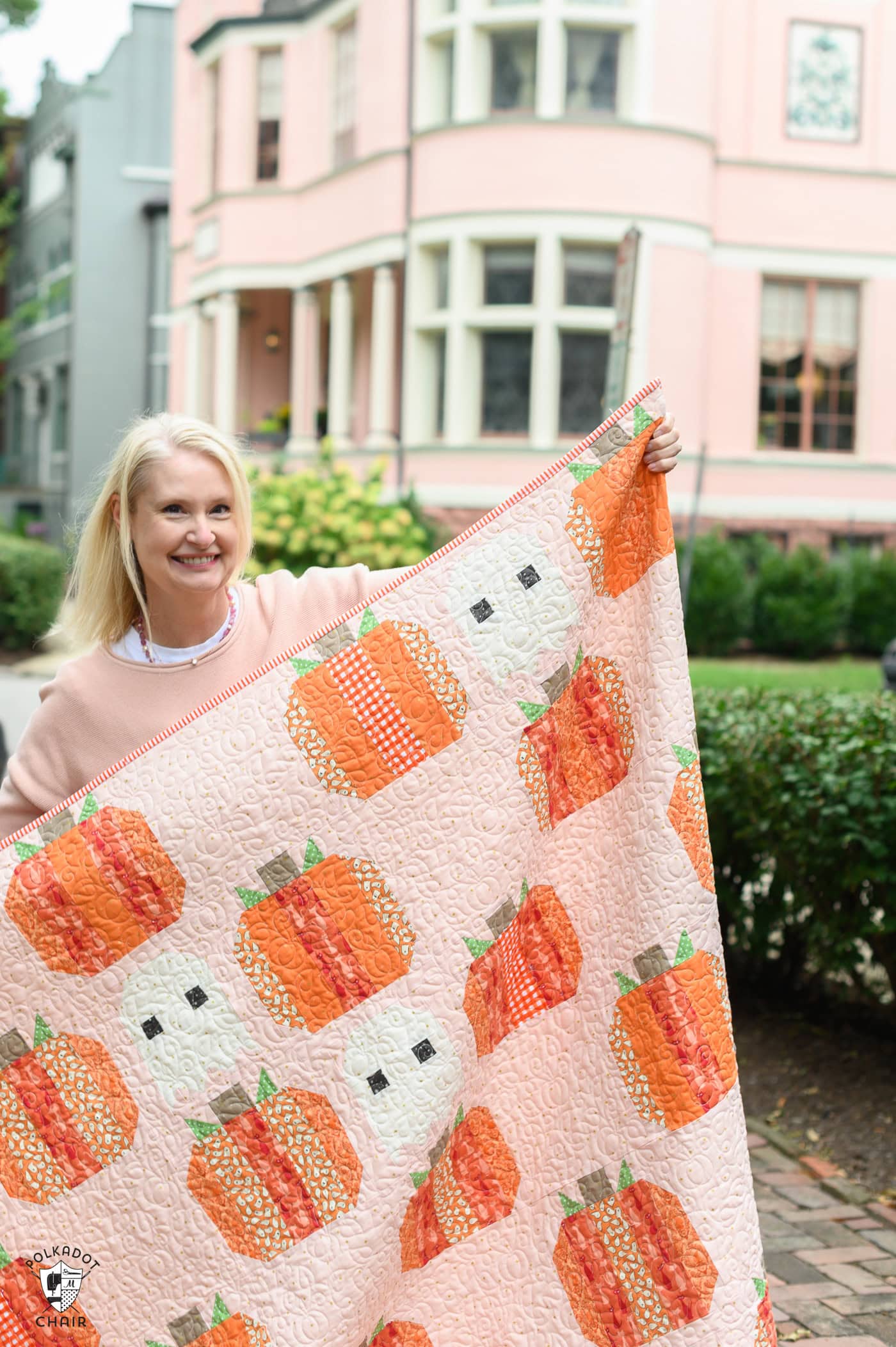 Woman holding a pumpkin-themed quilt outside a pink house.