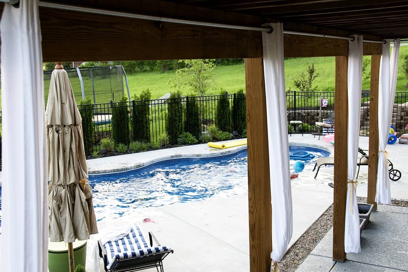 wood deck with attached white outdoor curtains and pool in background