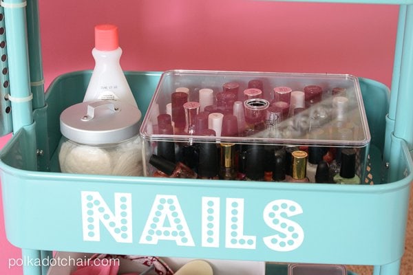 Blue shelf with the word NAILS in white vinyl filled with a clear bin of nail polish and a jar of cotton pads and a bottle of nail polish remover. 