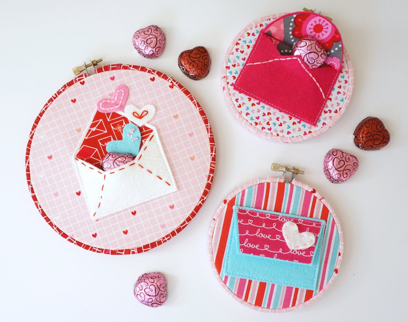 Multiple felt envelopes of embroidery hoops next to Valentines day chocolates.