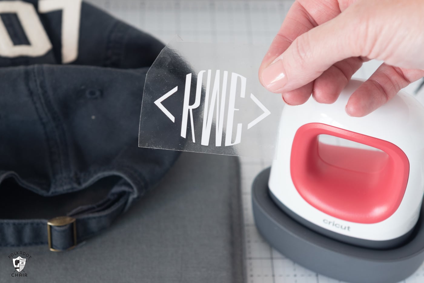 Cricut EasyPress mini, a navy baseball cap and a hand holding a sheet of iron on vinyl with a white monogram cut out "KME".