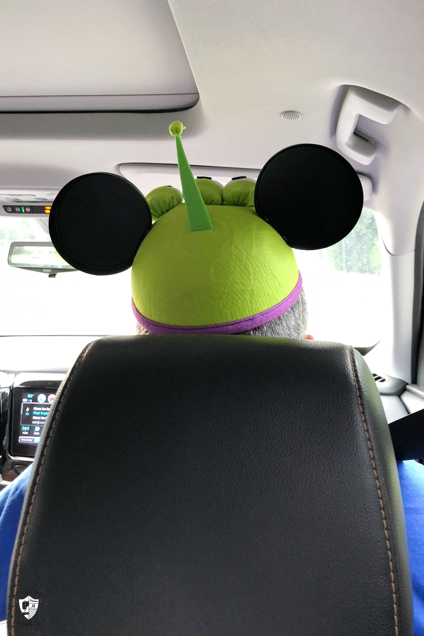 Sitting in a car taken from behind you see the back of the seat and the top of a mans head wearing a green alien mickey ear hat. 