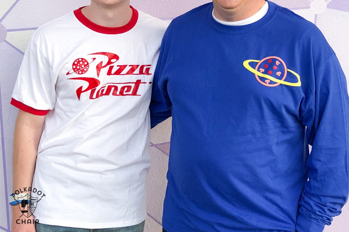 Zoomed in on two men's tshirts. One is white with red lining the collar and sleeve with the Pizza Planet logo. The other is in a dark blue long sleeve tshirt with a pizza surrounded by a ring in the top corner. 