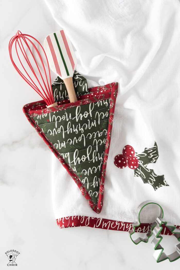 Christmas Tree potholder with whisk and spatula in pocket on table with kitchen towel. 