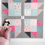 Pink, blue, gray & black baby quilt hanging on wall with pink chair and stuffed animals