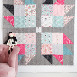 Pink, blue, gray & black baby quilt hanging on wall with pink chair and stuffed animals