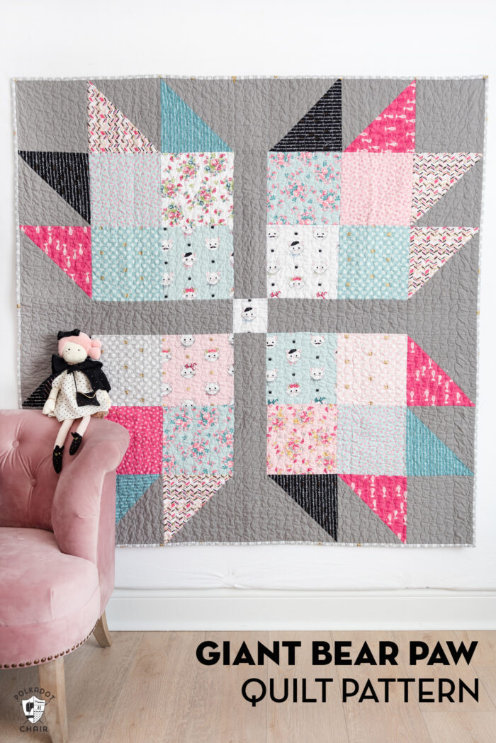 Pink, blue, gray & black baby quilt hanging on wall with pink chair and stuffed animals