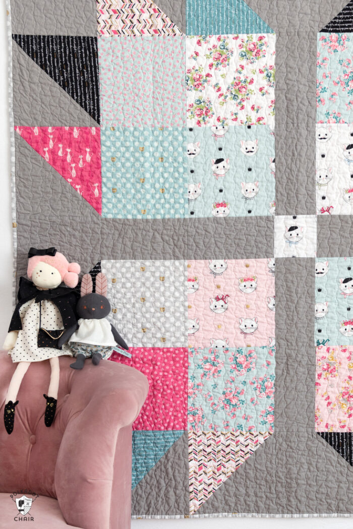 Pink, blue, gray & black baby quilt hanging on wall with pink chair and stuffed animals