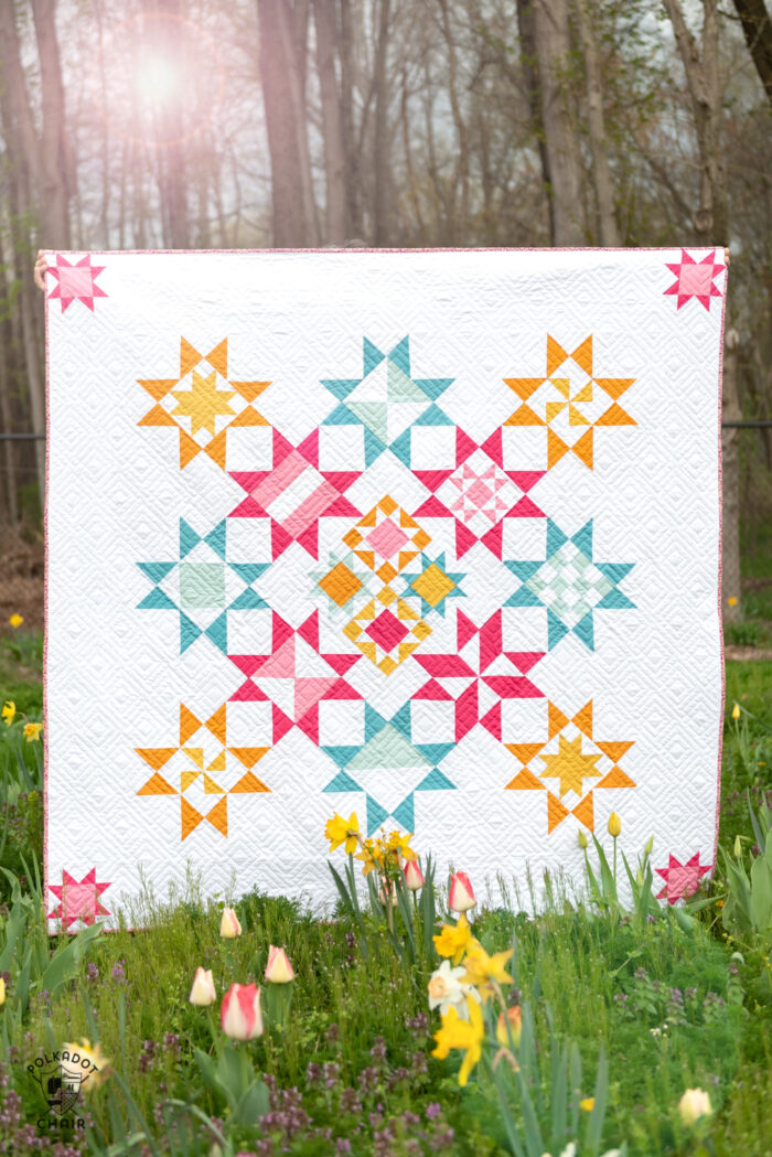 Brightly colored quilt with pink, blue and yellow stars in field of flowers. 