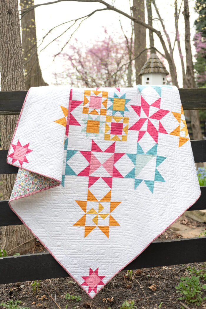 pink, blue and yellow geometric quilt in field of flowers hanging on fence