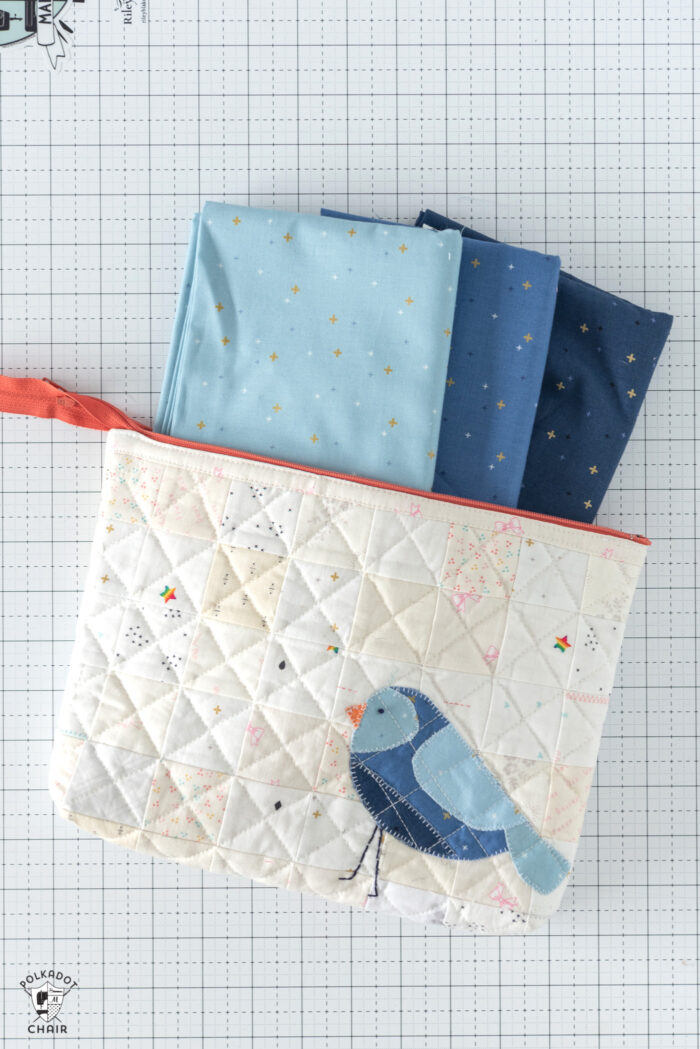 Ivory quilted zip pouch with blue bird on white countertop with blue fabrics poking out the top