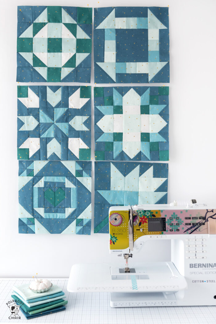 Six blue and aqua quilt blocks on white wall in front of sewing machine