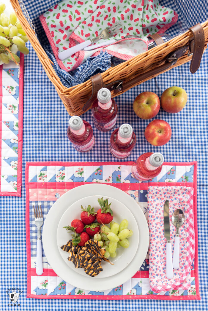 Outdoor table top with blue gingham tablecloth, food, picnic basket and quilted placemats