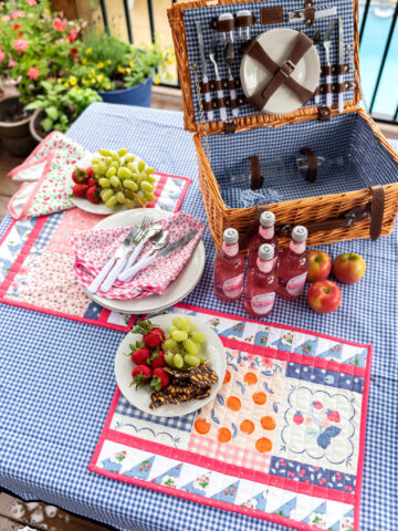 Outdoor table top with blue gingham tablecloth, food, picnic basket and quilted placemats