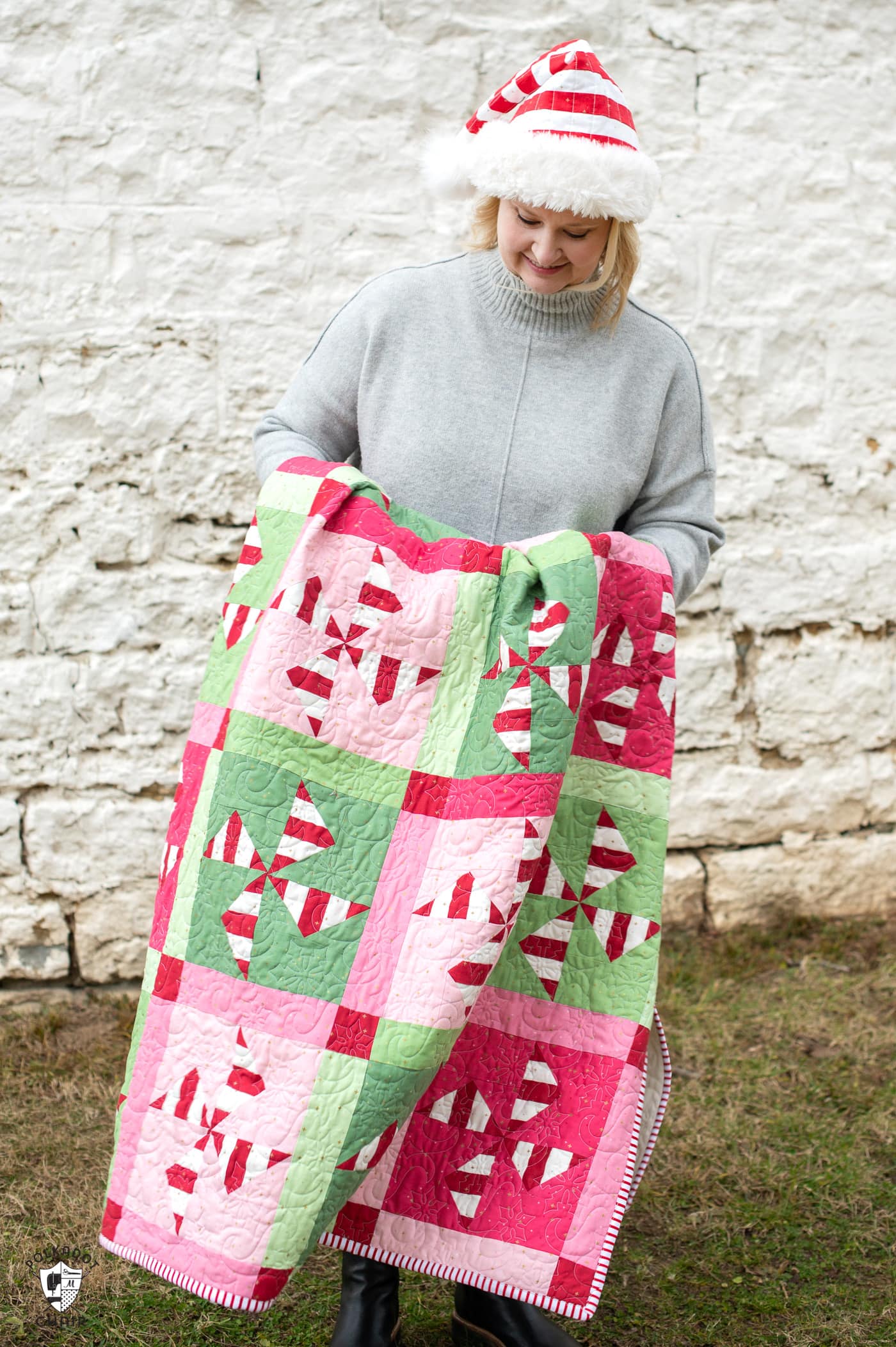 woman holding red, white, pink and green christmas quilt outdoors in front of a white brick wall