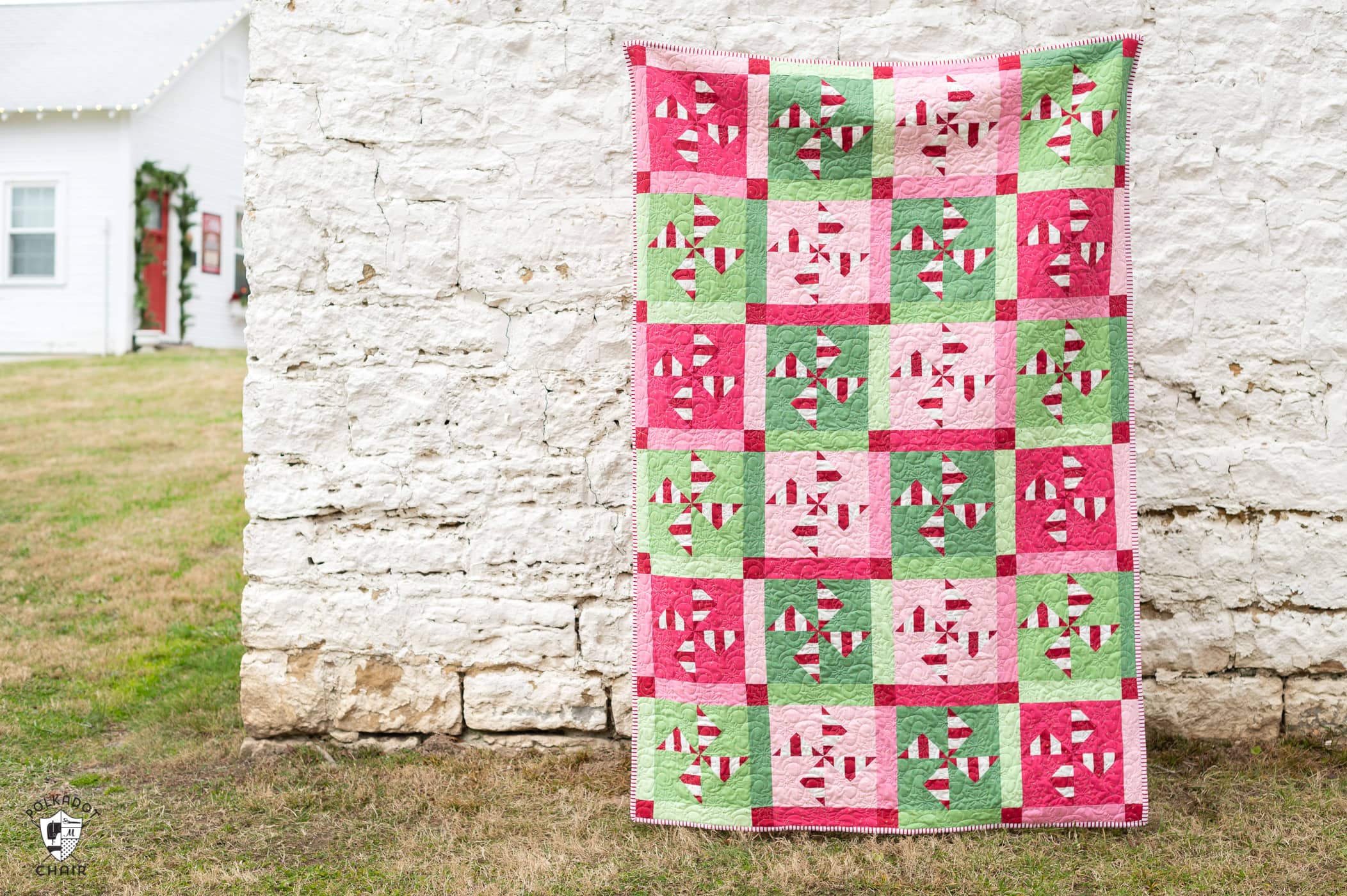 woman holding red, white, pink and green christmas quilt outdoors in front of a white brick wall