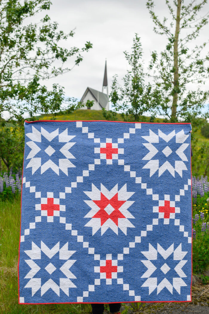 blue and red quilt outdoors with church and flowers in background