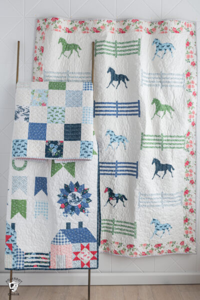 Three qulits one on wall and two on quilt rack, white with blue, green and pink fabric and horse motifs