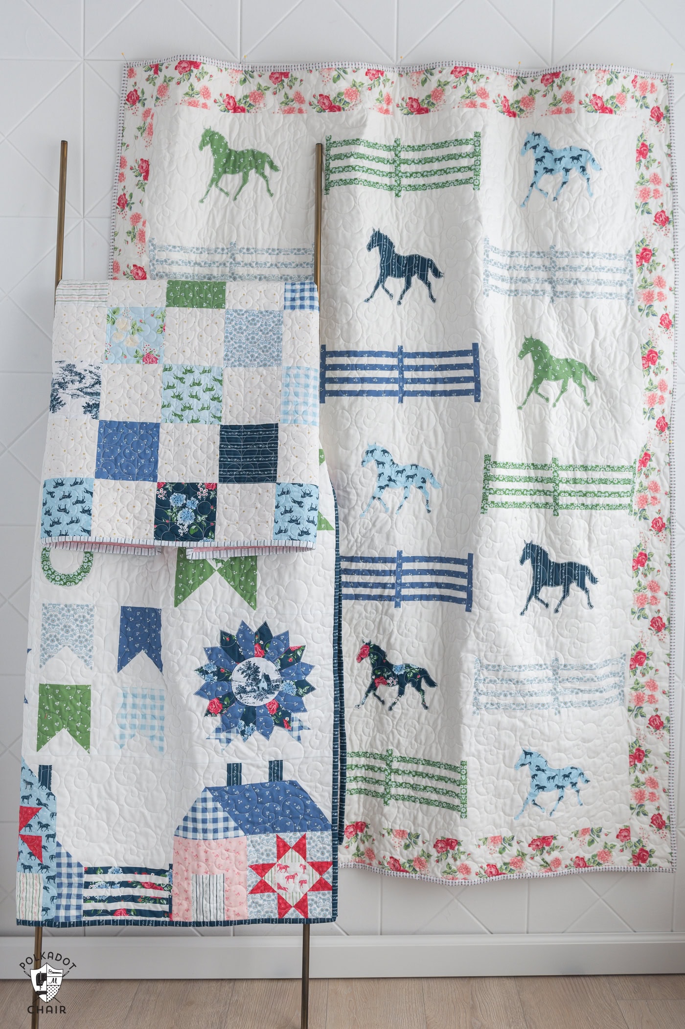 Three qulits one on wall and two on quilt rack, white with blue, green and pink fabric and horse motifs