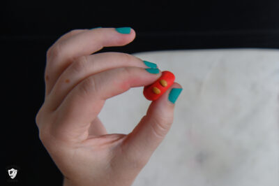 hand with teal nails, holding flattened red clay bead with two small gold dots.