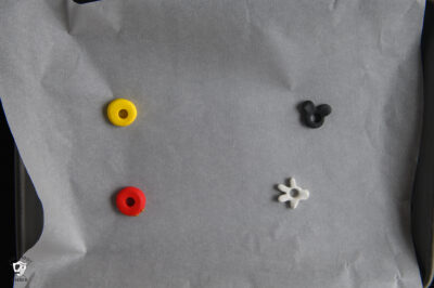 baking sheet with yellow round clay bead with hole in the middle, black mickey head clay bead with hole in the middle, red round clay bead with hole in the middle, and white glove shaped clay bead with clay in the middle.