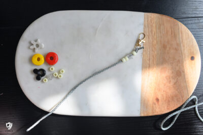 lobster clasp with gray paracord, with white and clear beads on a marble slab with red, yellow, and black beads beside.