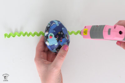 blue egg with green ricrac underneath with a pink hot glue gun putting glue on the ricrac.