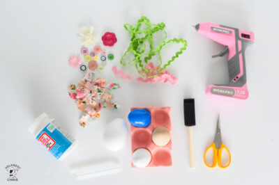 pink egg carton with a blue, white, and brown faux egg. green ricrac. pink hot glue gun. cut up pieced of pink fabric. yellow scissors. black foam paint brush. modgepodge bottle.