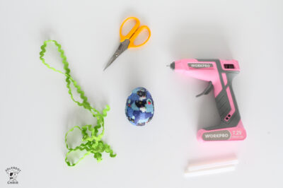 blue egg, green ricrac, pink hot glue gun, and yellow scissors on a white table.