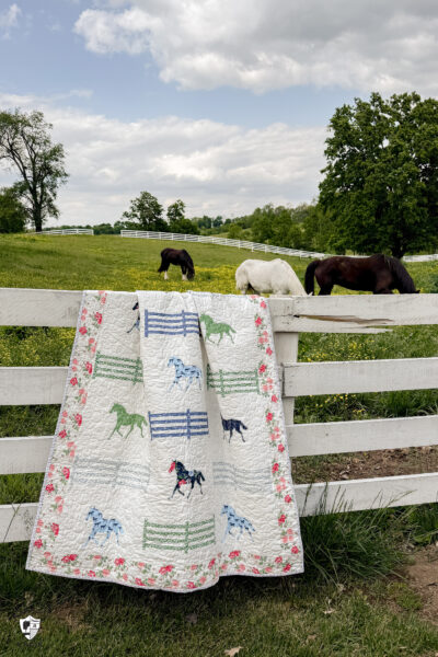 horse quilt hanging on white horse fence in front of field with horses and yellow flowers