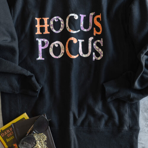 Black sweatshirt with pink, orange and purple hocus pocus lettering on gray table